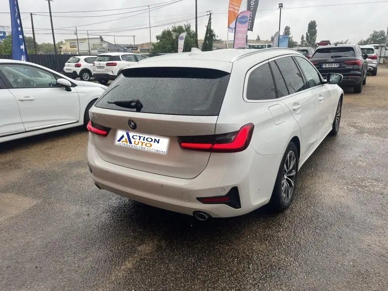BMW Série 3 316dA Business Design Alpinweiss vue 3/4 arrière droit sous la pluie avec feux arrière LED allumés.