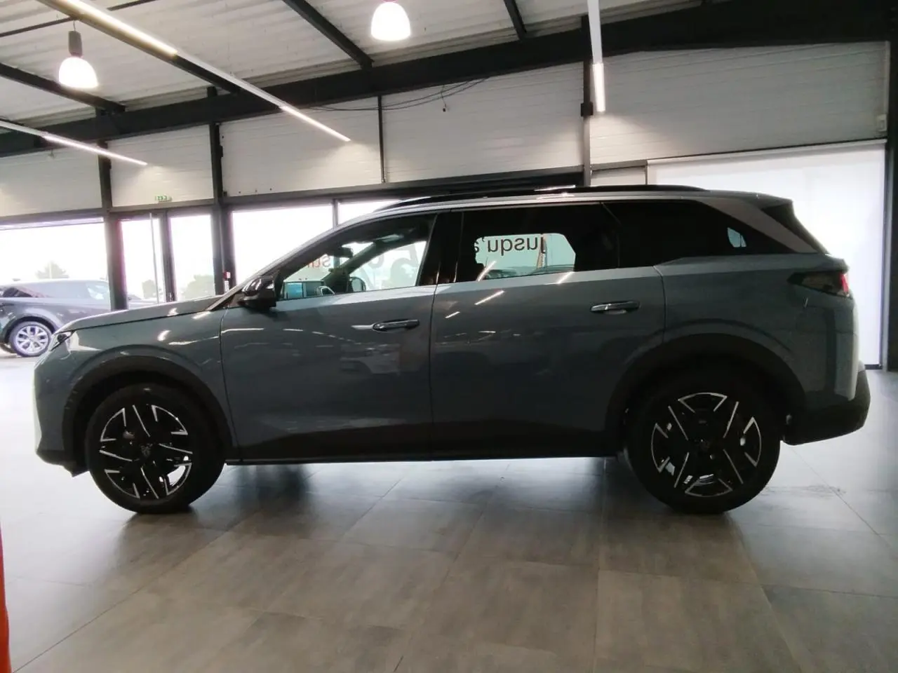 Vue de profil côté gauche du Peugeot 5008 2025 bleu Ingaro avec jantes alliage noires diamantées en intérieur showroom.