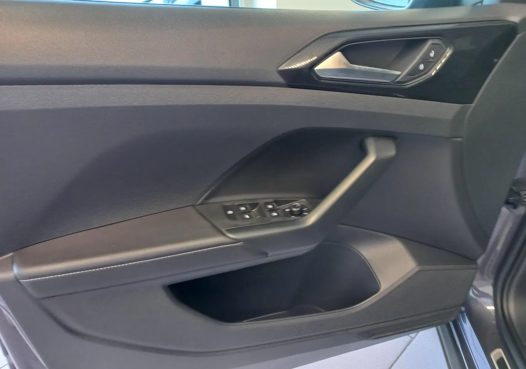 Vue intérieure côté gauche de la porte avant d'un Volkswagen T-Cross gris cendré avec commandes électriques des vitres.