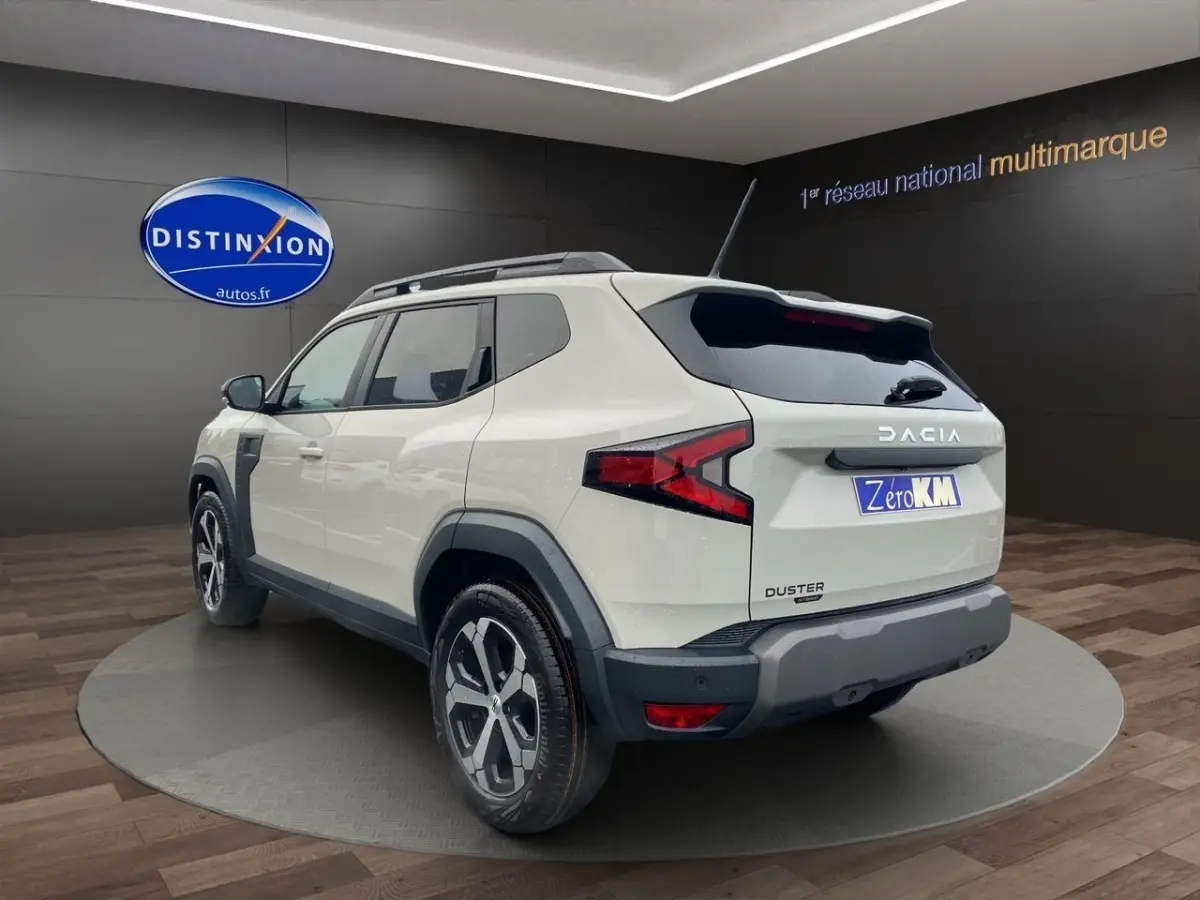Vue 3/4 arrière droite du Dacia Duster beige 2025 avec barres de toit noires et jantes alliage visibles.