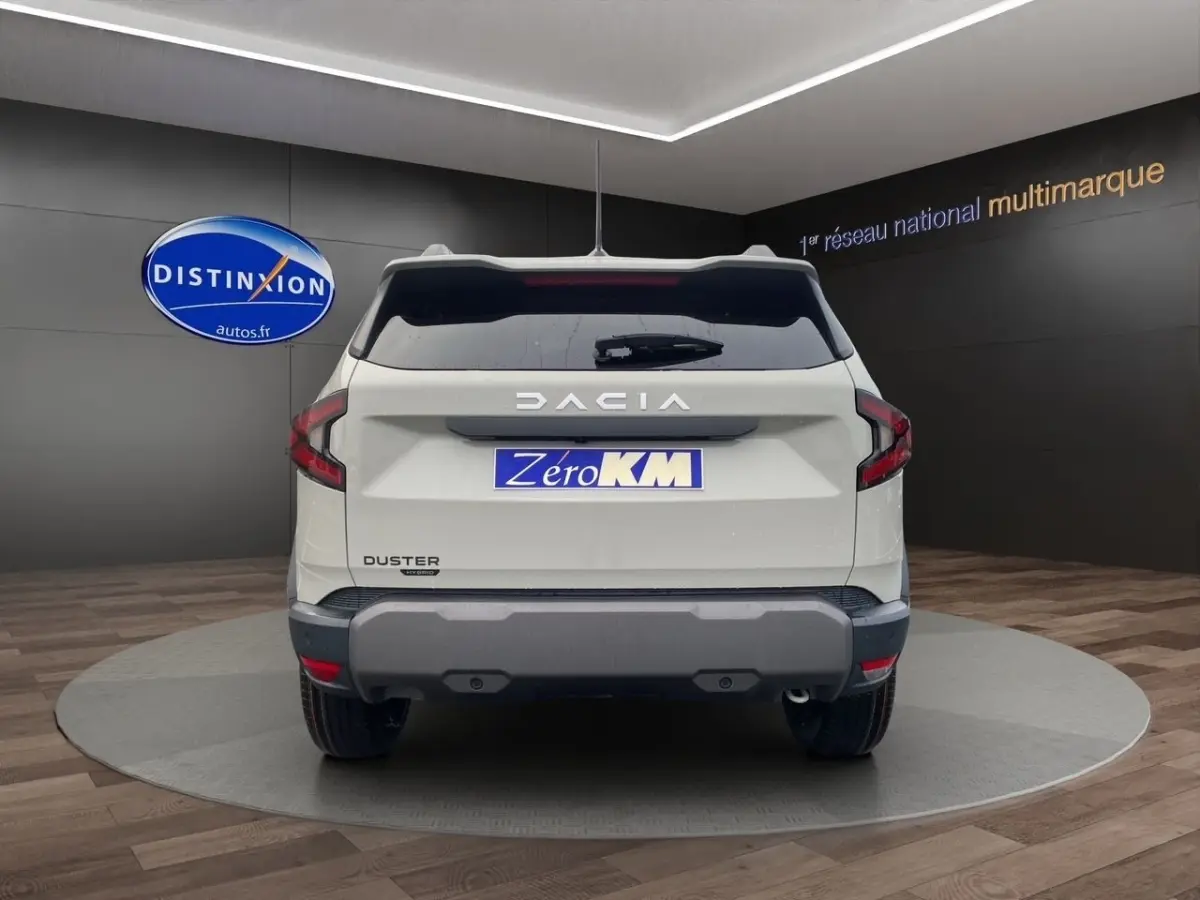 Vue arrière d'un Dacia Duster beige 2025 avec logo DACIA lumineux et plaque Zero KM dans un showroom.