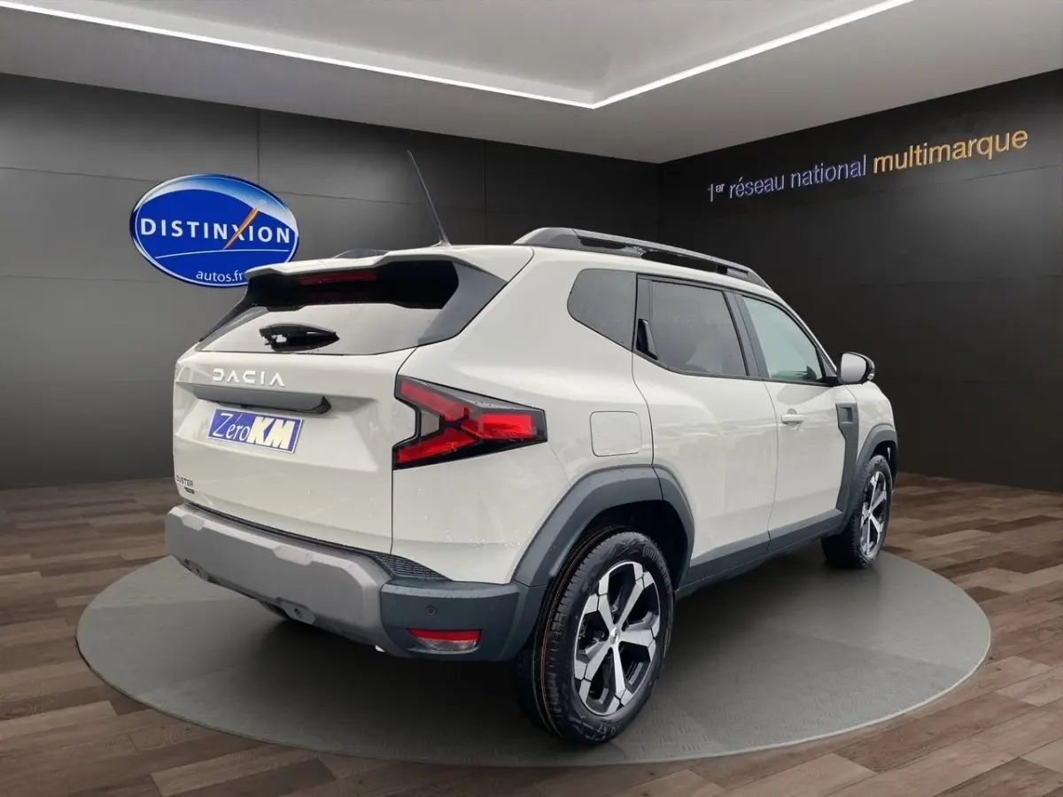 Vue 3/4 arrière droite du Dacia Duster beige 2025 avec jantes alliage et barres de toit noires.