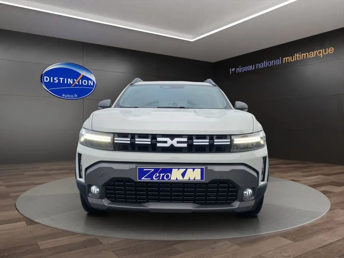 Vue avant d'un Dacia Duster beige 2025 avec calandre noire et phares LED allumés en showroom.