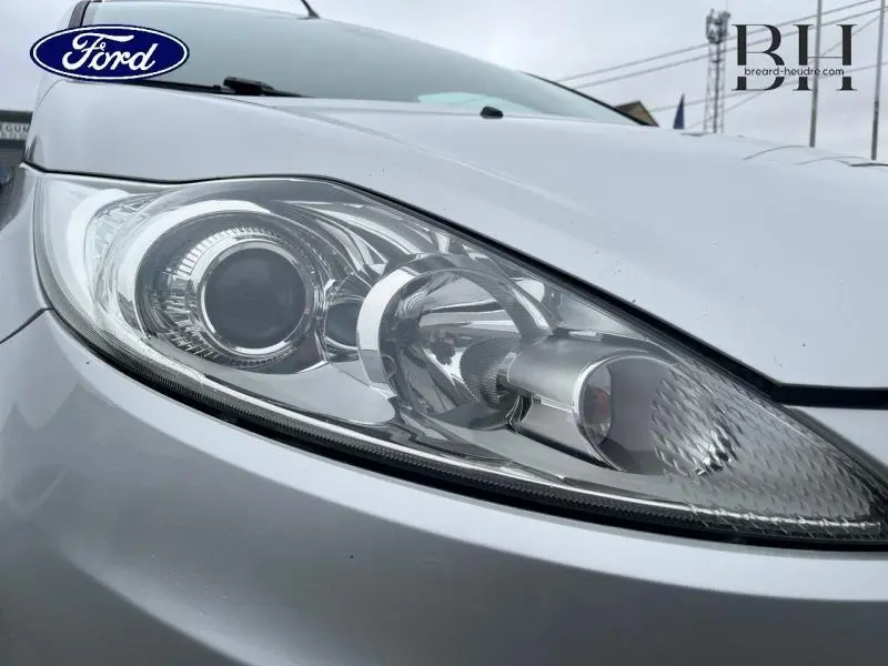 Gros plan sur le phare avant gauche gris lunaire de la Ford Fiesta 2012, soulignant ses détails lumineux et la carrosserie lisse.