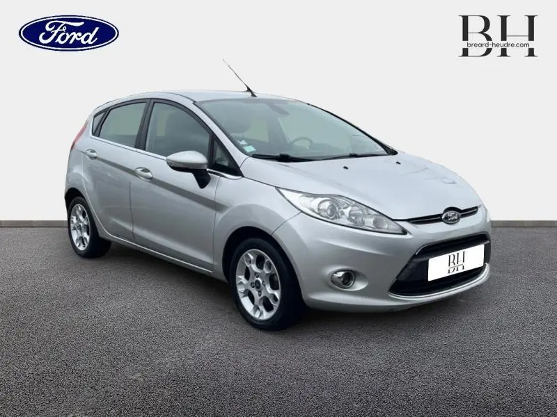 Vue 3/4 avant d'une Ford Fiesta gris lunaire de 2012 avec jantes alu et calandre chromée sur fond neutre.
