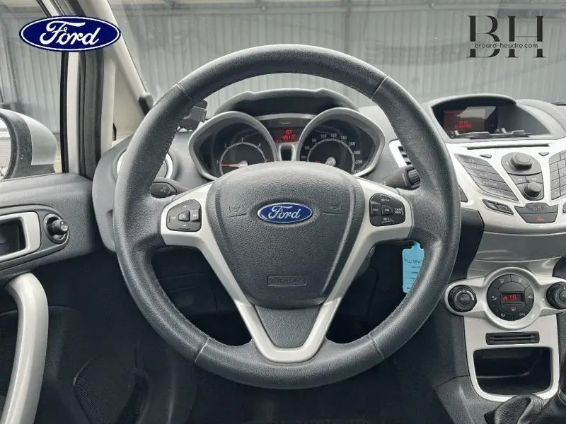 Vue intérieure centrée sur le volant cuir noir avec commandes audio d'une Ford Fiesta gris lunaire de 2012.