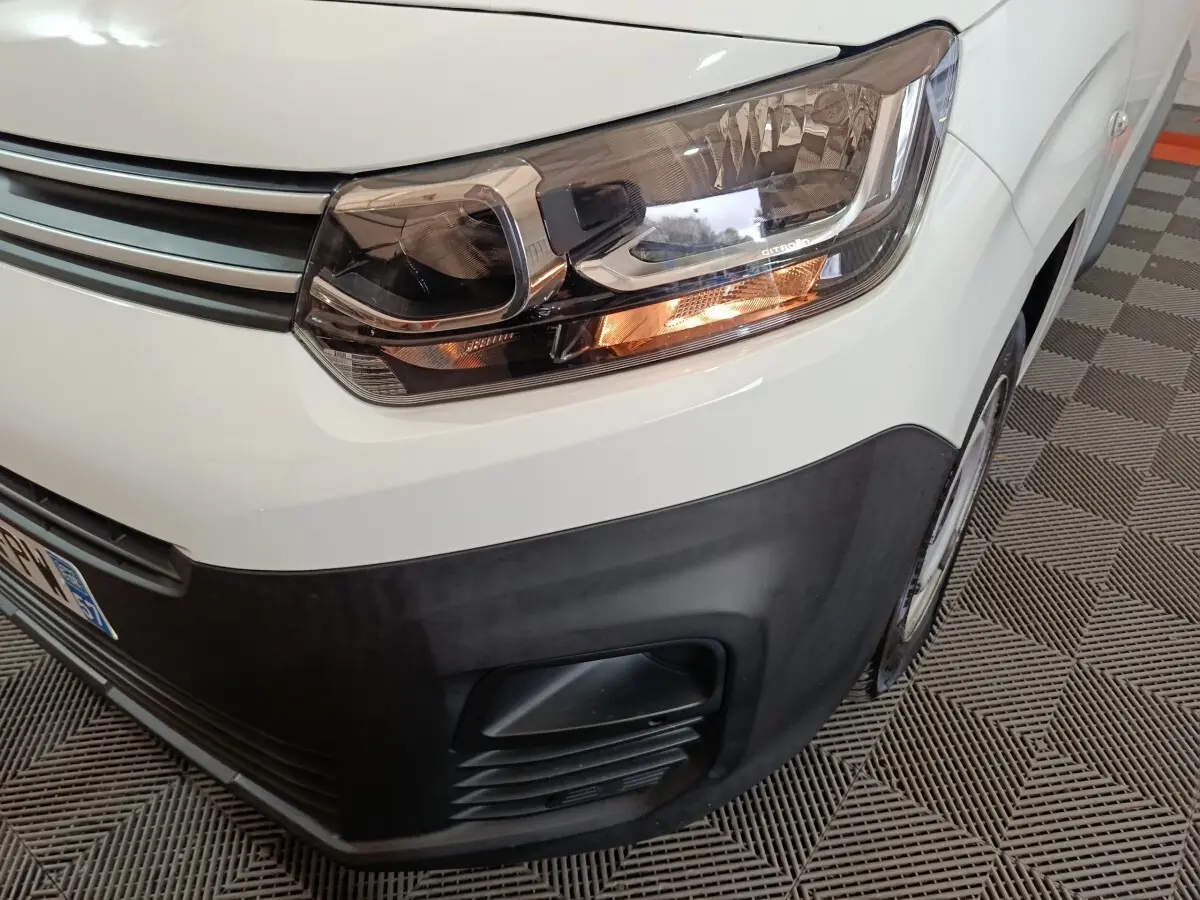 Gros plan sur le phare avant droit et la calandre noire d’un Citroën Berlingo blanc, vue en légère plongée.
