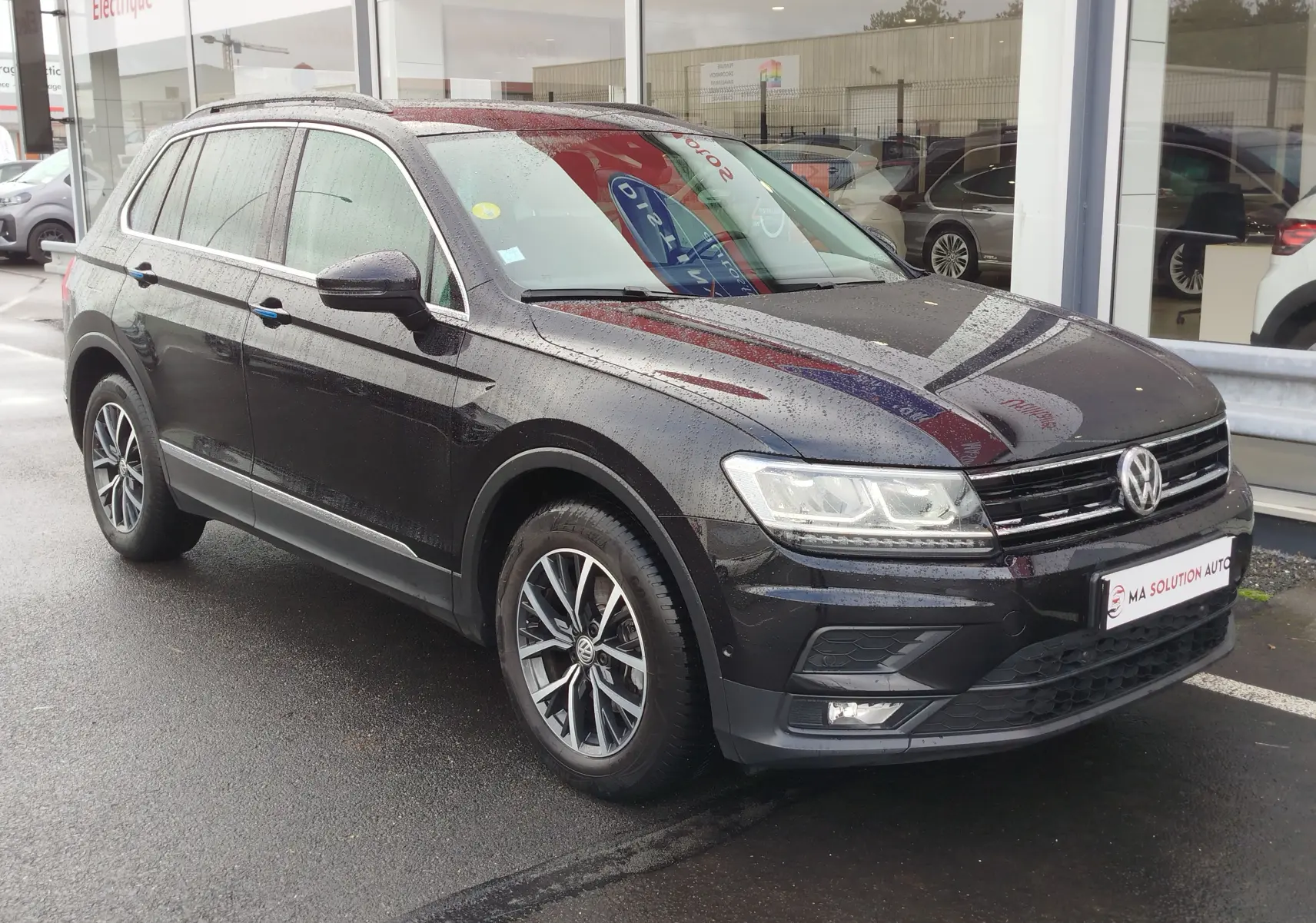 Volkswagen Tiguan 2.0 TDI noir nacré vu en 3/4 avant droit sous la pluie, avec jantes alliage et feux LED.