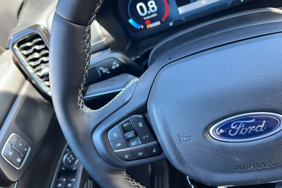 Gros plan sur le volant cuir multifonctions noir avec logo Ford et commandes intégrées, tableau de bord numérique en arrière-plan.