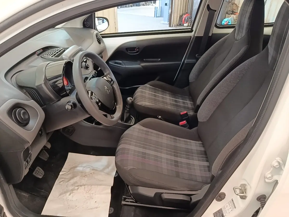Vue intérieure côté conducteur de la Peugeot 108 blanche, montrant les sièges avant à motifs carreaux et le volant multifonctions.