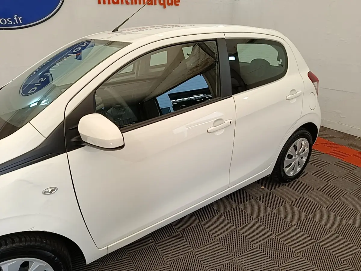 Vue de profil côté gauche d'une Peugeot 108 blanche stationnée en intérieur sur sol à motifs.
