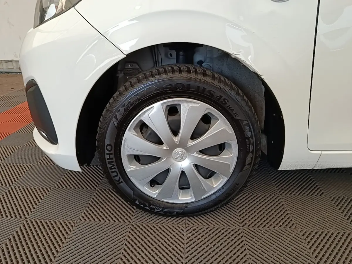 Gros plan sur la roue avant gauche d'une Peugeot 108 blanche, avec enjoliveur argenté et carrosserie légèrement froissée.