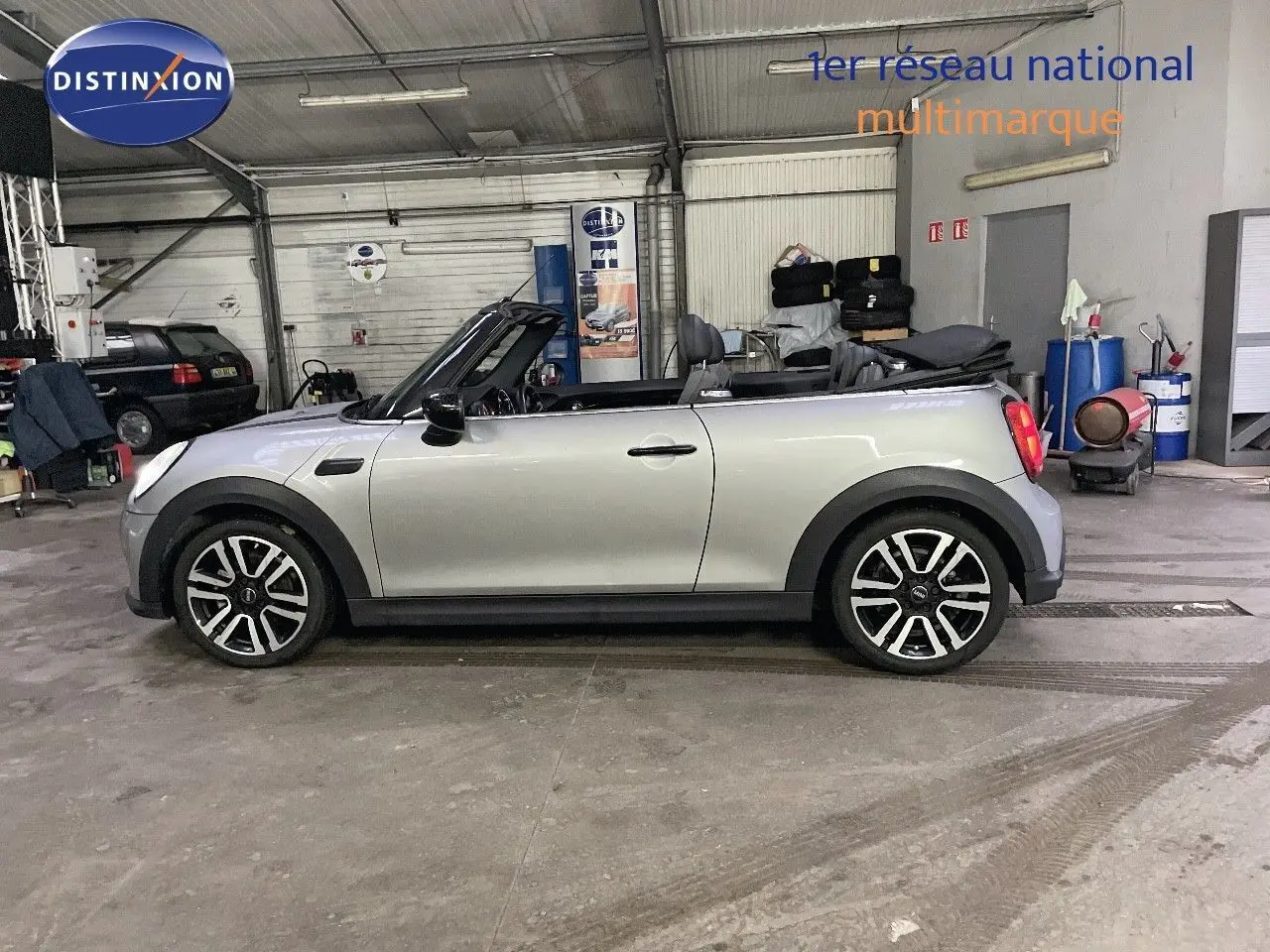 Vue de profil côté gauche d'une MINI Cabriolet F57 2023 Melting Silver Metal avec capote ouverte en intérieur d'atelier.