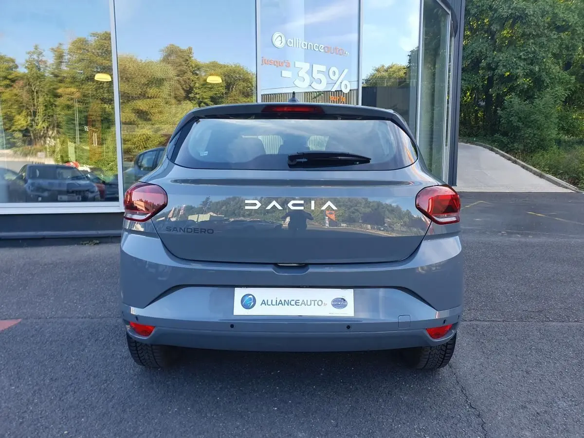 Vue arrière d'une Dacia Sandero 2024 gris urbain avec feux arrière rouges et logo DACIA visible.