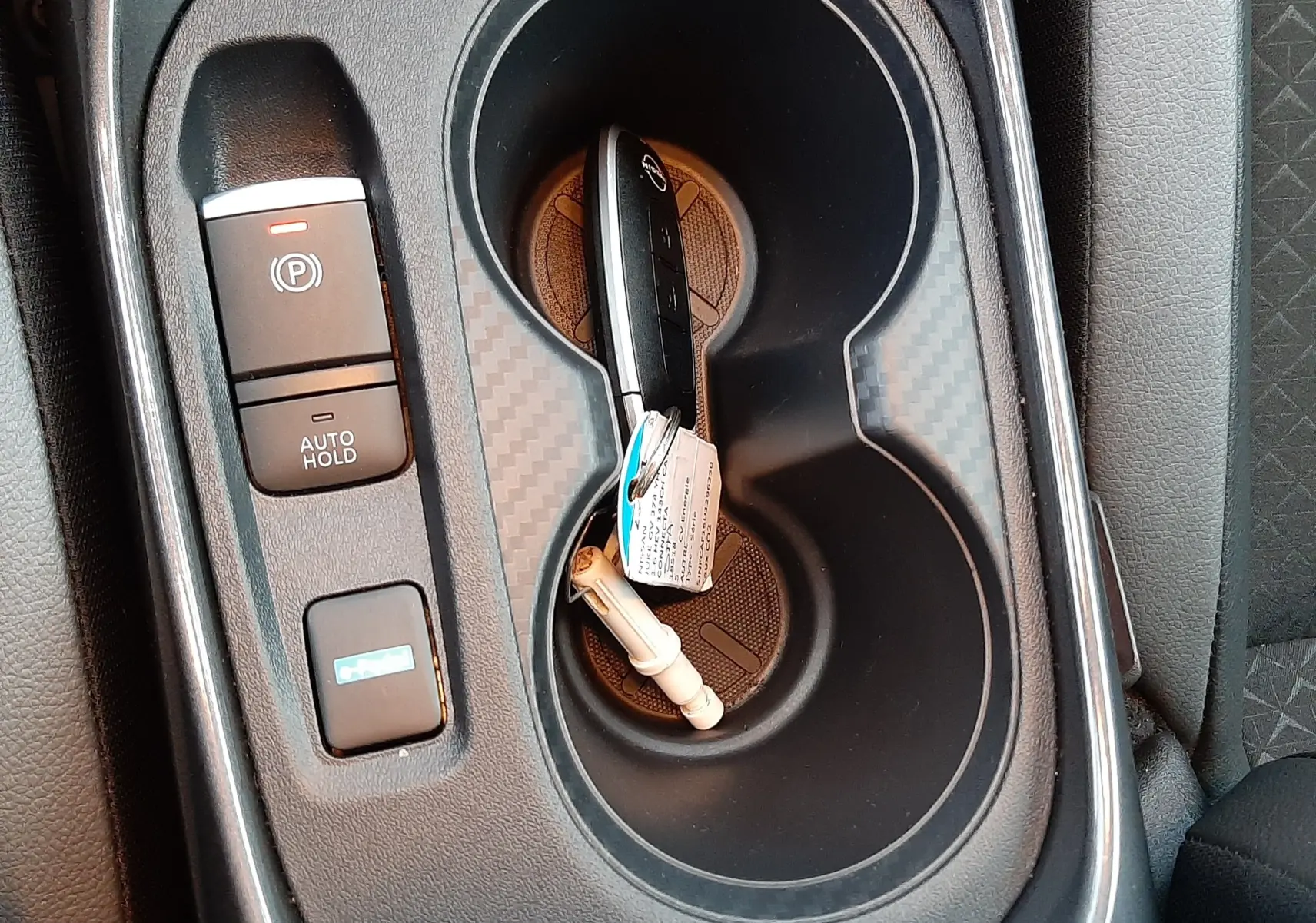 Gros plan sur la console centrale noire du Nissan Juke 2024 avec clé hybride dans porte-gobelets et bouton frein à main électrique.