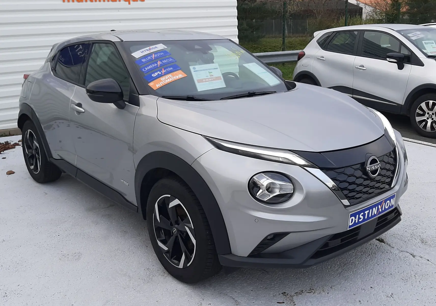 Nissan Juke 1.6 HEV gris perle métal vu en 3/4 avant droit, avec jantes noires et stickers promotionnels sur le pare-brise.
