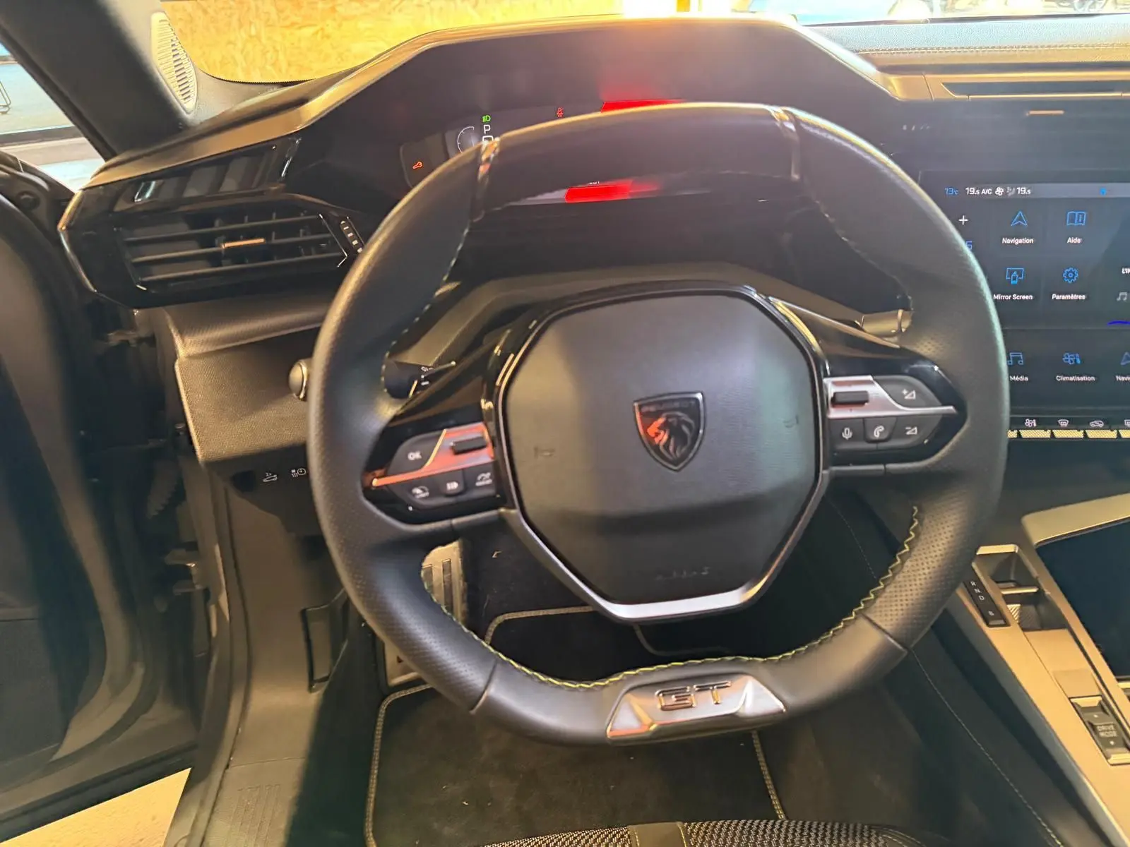 Vue intérieure centrée sur le volant noir perforé avec logo Peugeot et badge GT, planche de bord et écran tactile visibles.