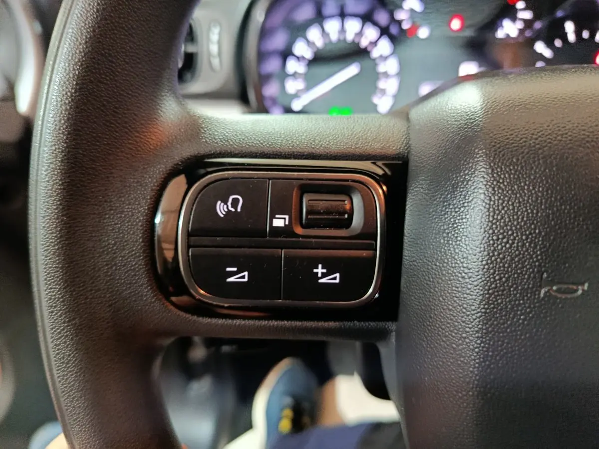 Gros plan sur les commandes gauche du volant multifonctions noir du Citroën C3 2024, avec tableau de bord flou en arrière-plan.