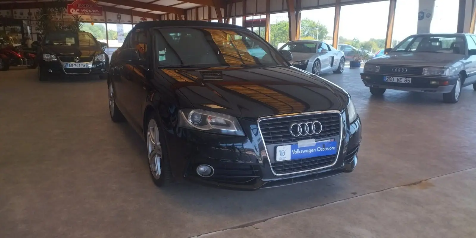 Audi A3 Cabriolet noir vue 3/4 avant droit dans un showroom avec plusieurs autres véhicules exposés.