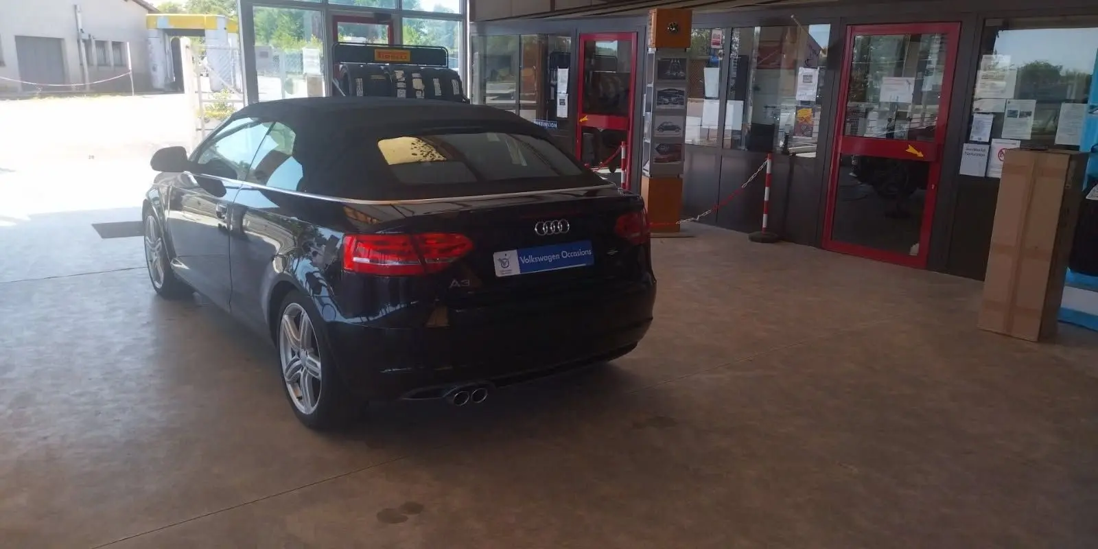 Audi A3 Cabriolet noir vue 3/4 arrière droit, capote fermée, garée dans un garage lumineux avec portes vitrées rouges.