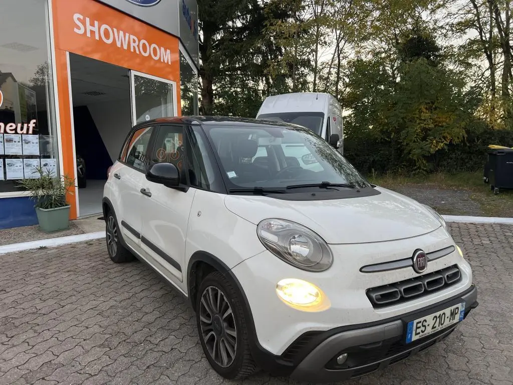 Vue 3/4 avant droite d'une Fiat 500L blanche avec toit noir bi-color, feux allumés, devant un showroom.