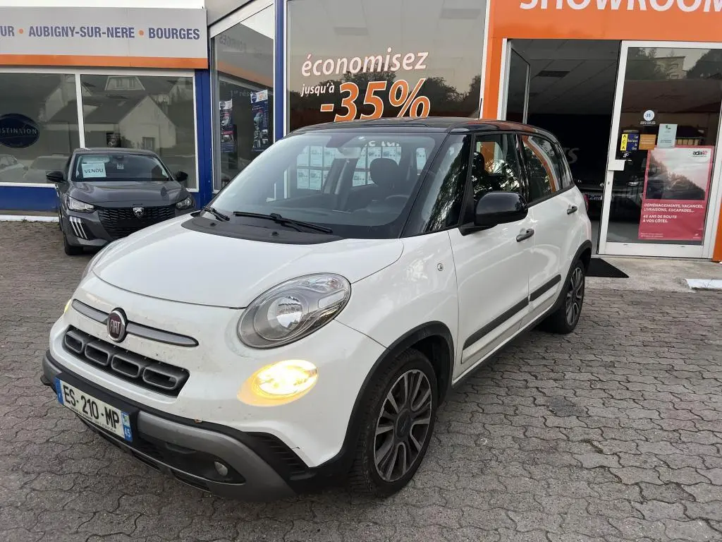 Vue 3/4 avant d'une Fiat 500L blanche avec toit noir et feux diurnes allumés devant un showroom.