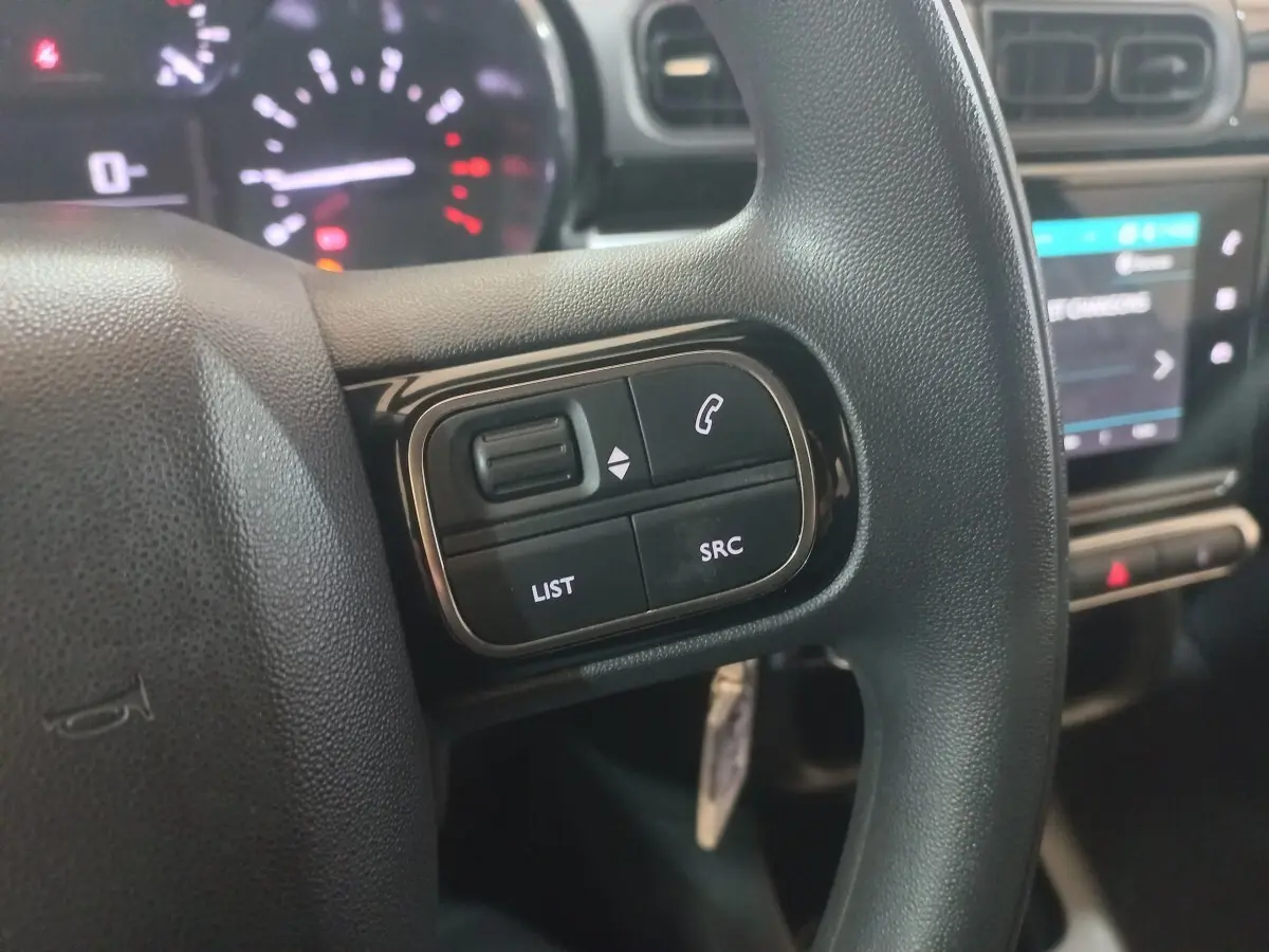 Gros plan sur les commandes gauche du volant noir de la Citroën C3 2024 avec tableau de bord et écran tactile en arrière-plan.