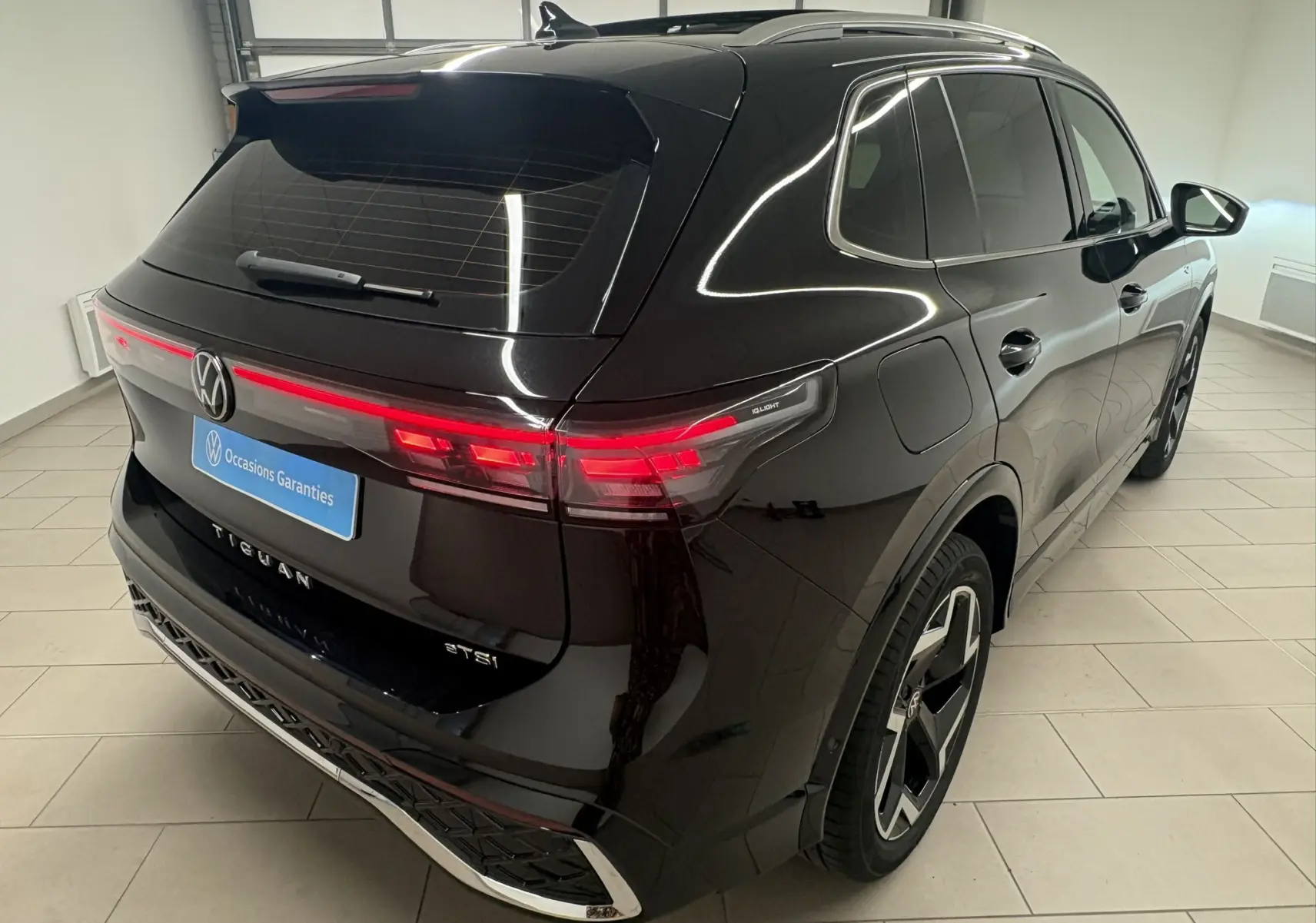 Vue 3/4 arrière droite du Volkswagen Tiguan 2025 noir intense nacré avec feux arrière allumés et toit ouvrant visible.