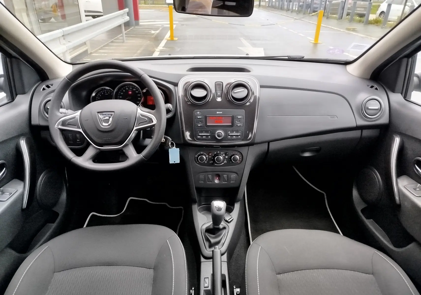 Vue intérieure frontale du tableau de bord et volant noir de la Dacia Sandero 2019 avec commandes centrales et sièges tissu gris.