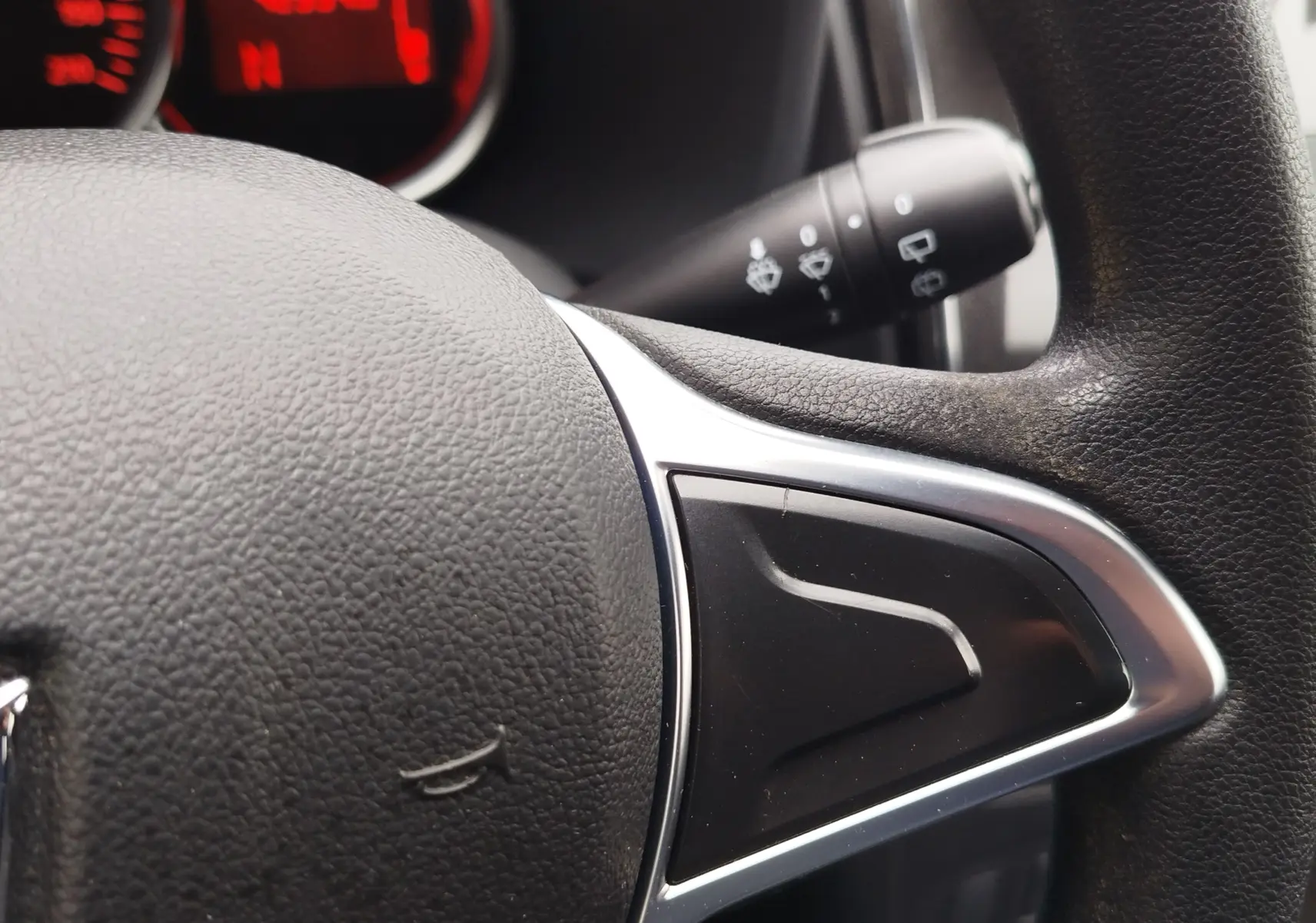 Gros plan sur le volant en cuir noir de la Dacia Sandero blanc glacier, avec commandes intégrées et tableau de bord rouge en arrière-plan.