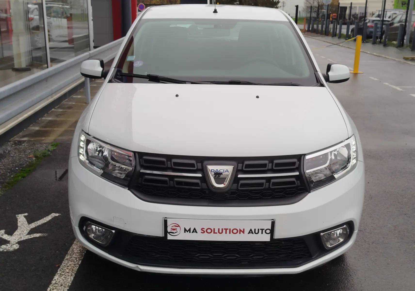 Vue frontale d'une Dacia Sandero blanc glacier de 2019 avec calandre noire et feux LED allumés.