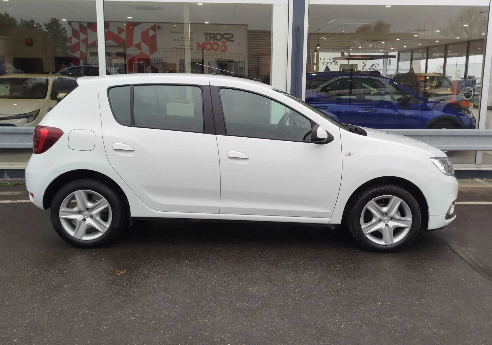Profil latéral droit d'une Dacia Sandero blanc glacier de 2019, compacte et cinq portes, stationnée devant une vitrine.