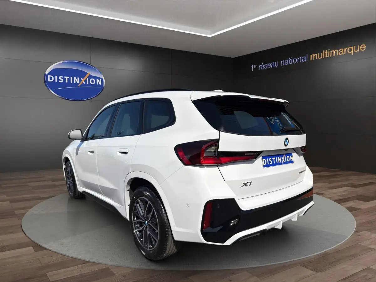 BMW X1 sDrive20d blanc vue 3/4 arrière droit, avec jantes noires et aileron de toit visible.
