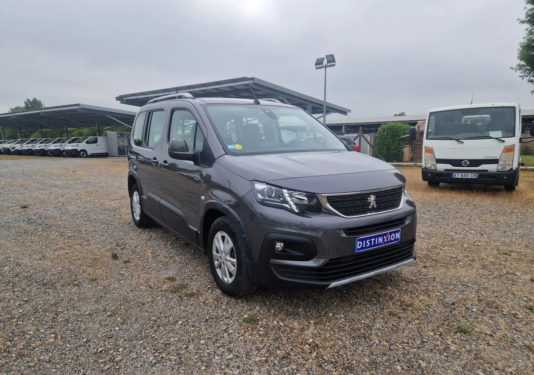 Peugeot Rifter gris platinium en 3/4 avant droit sur un sol gravillonné avec plusieurs véhicules en arrière-plan.