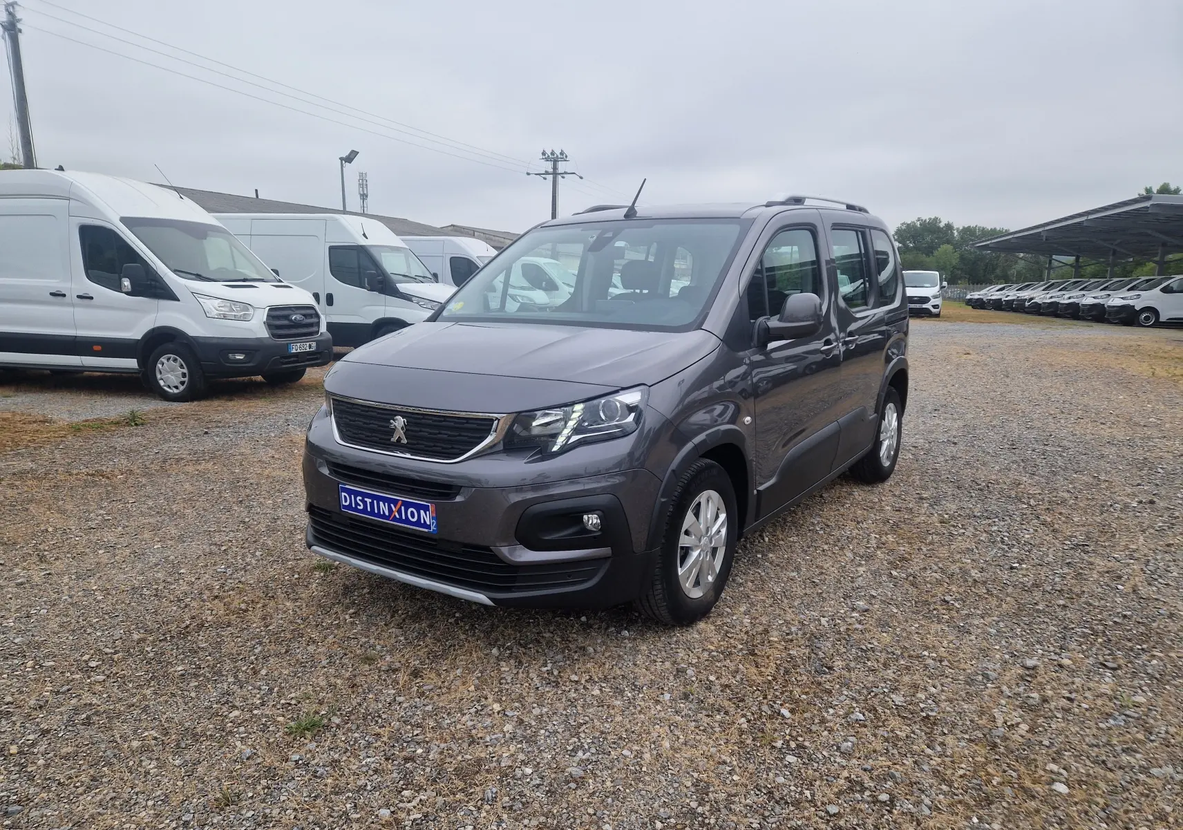 Peugeot Rifter gris platinium en 3/4 avant droit sur un parking gravillonné avec plusieurs fourgons blancs en arrière-plan.