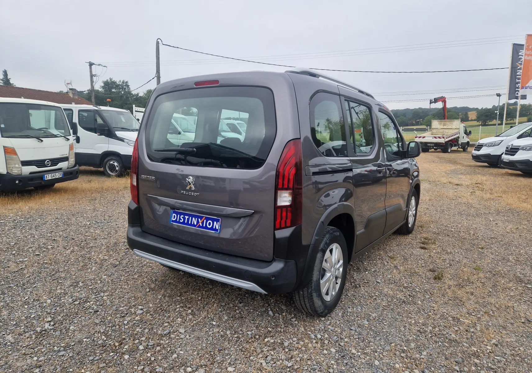 Vue 3/4 arrière droite d'un Peugeot Rifter gris platinium avec hayon et feux arrière verticaux, stationné sur un sol gravillonné.
