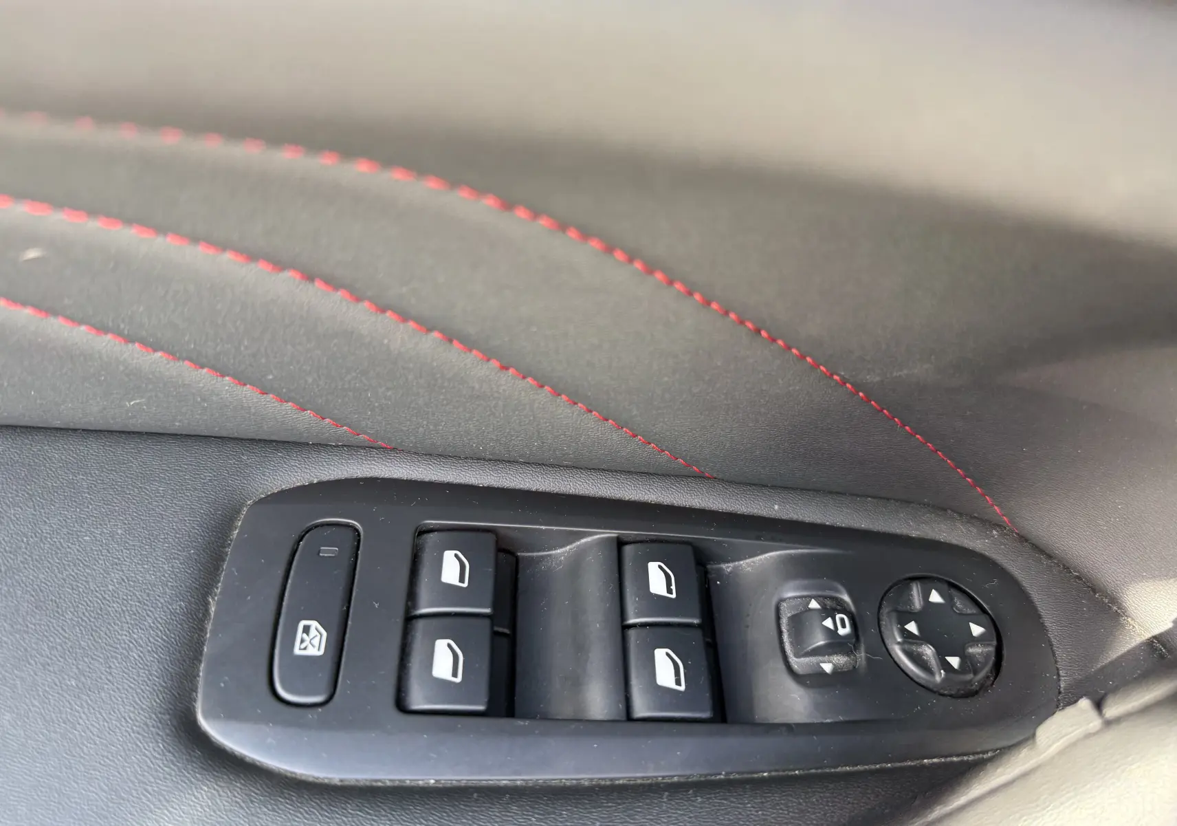 Gros plan sur les commandes des vitres électriques et réglages des rétroviseurs côté gauche, avec surpiqûres rouges sur la garniture noire.