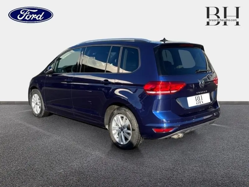 Vue 3/4 arrière droite d'un Volkswagen Touran bleu Atlantique métallisé avec barres de toit et jantes alu.