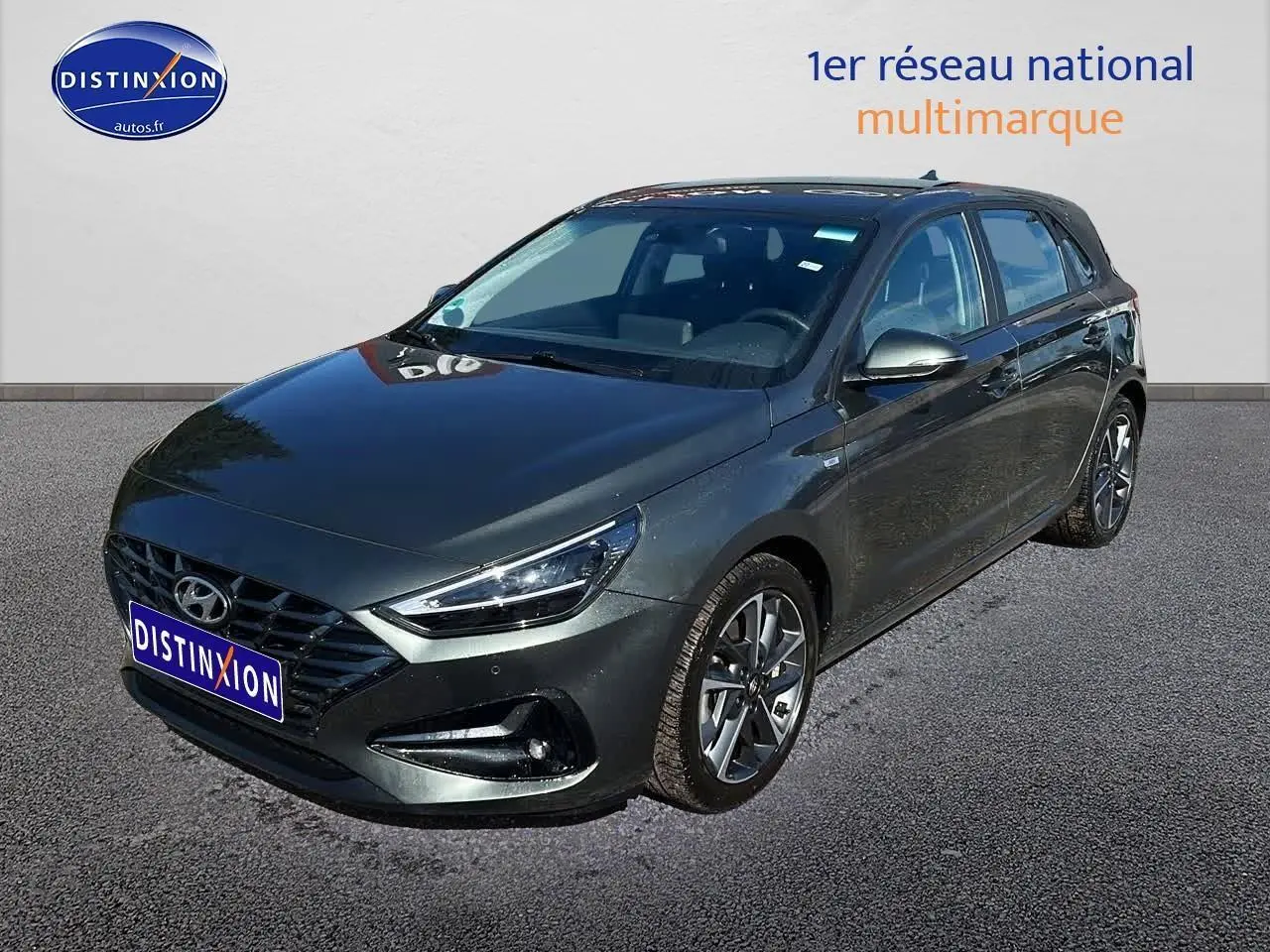Hyundai i30 gris argent vue 3/4 avant droit avec jantes alu et phares LED allumés sur parking.