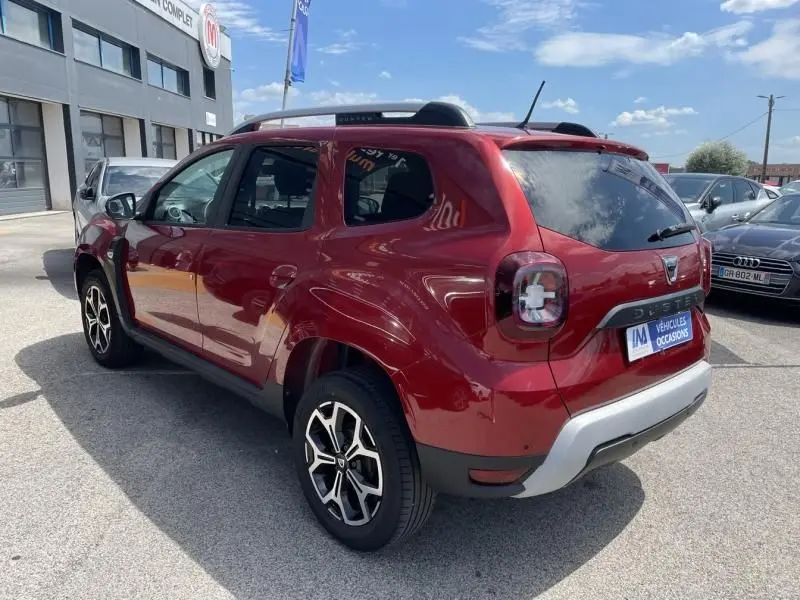 Vue 3/4 arrière droite d'un Dacia Duster TCE 150 Prestige 4x2 rouge métal avec jantes alliage et barres de toit noires.