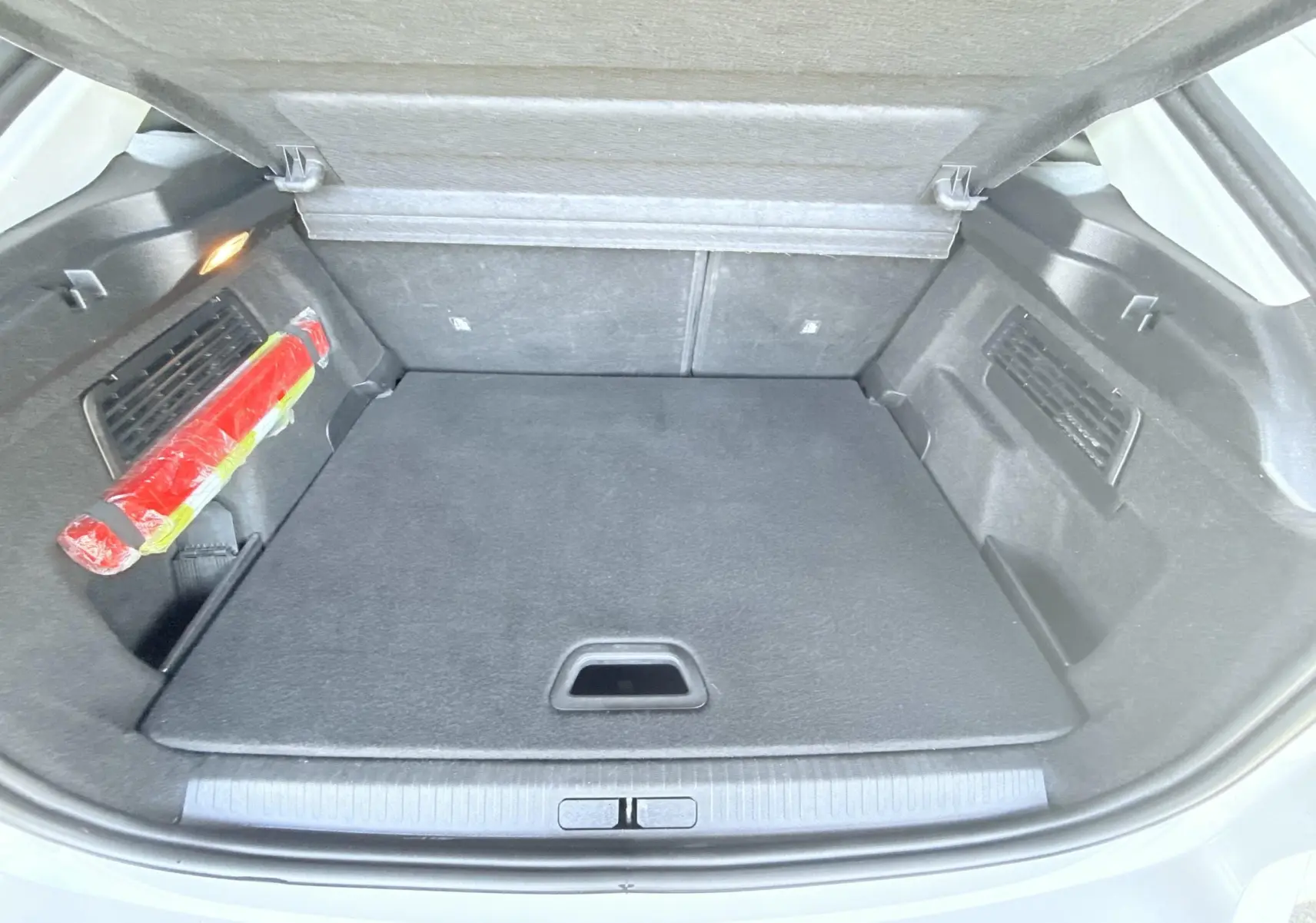 Coffre ouvert vu de l'arrière d'une Citroën C4 blanche, avec tapis de sol noir et triangle de signalisation rouge rangé sur le côté.