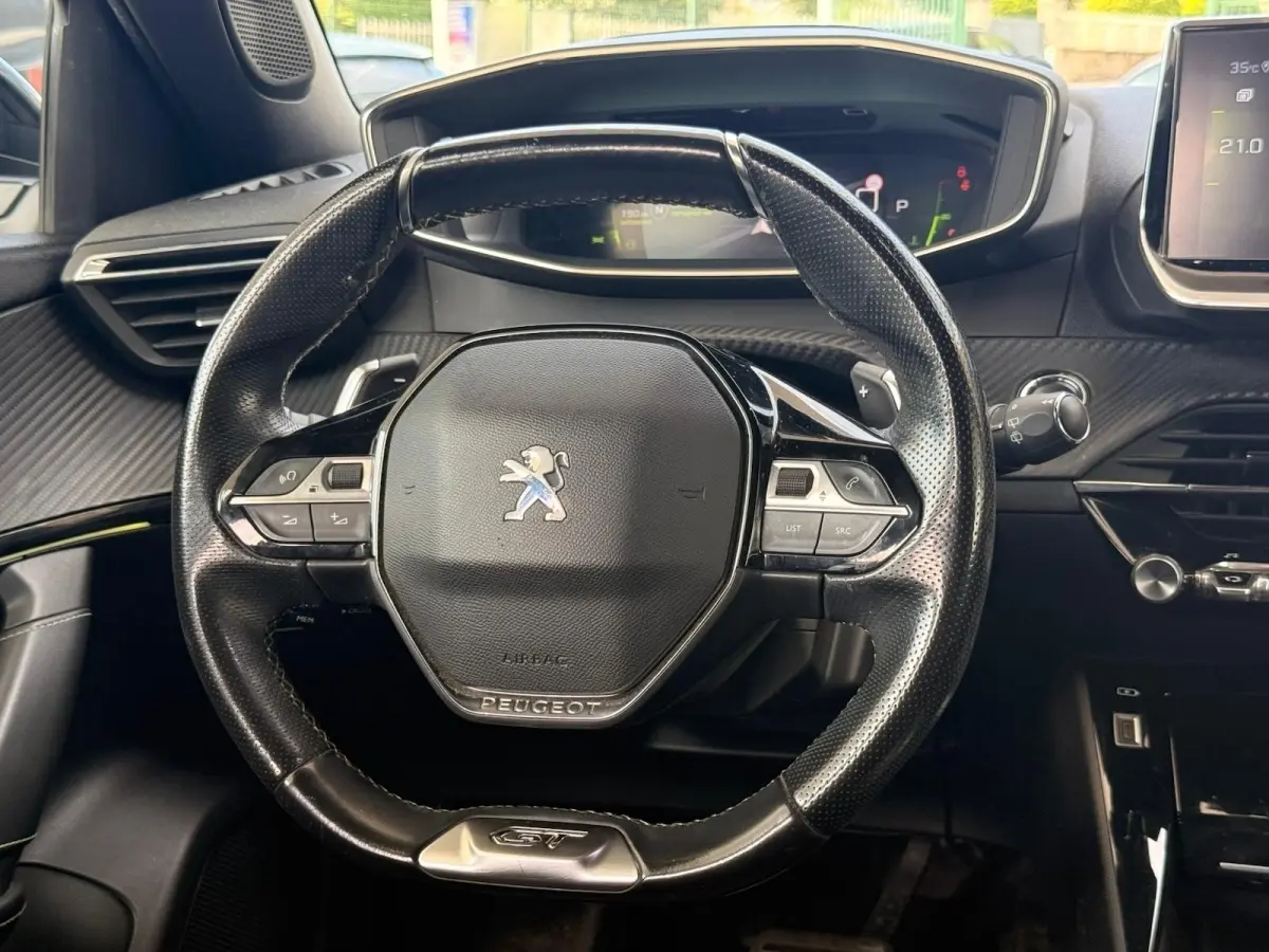 Vue rapprochée du volant noir perforé avec logo Peugeot et badge GT, tableau de bord et écran digital en arrière-plan.