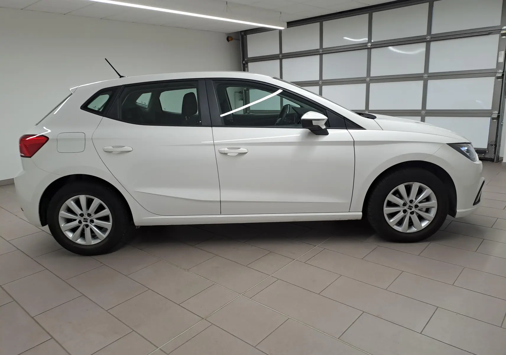 Vue de profil côté gauche d'une SEAT Ibiza blanche Candy 2023, finition Style Business, en intérieur sur sol carrelé.