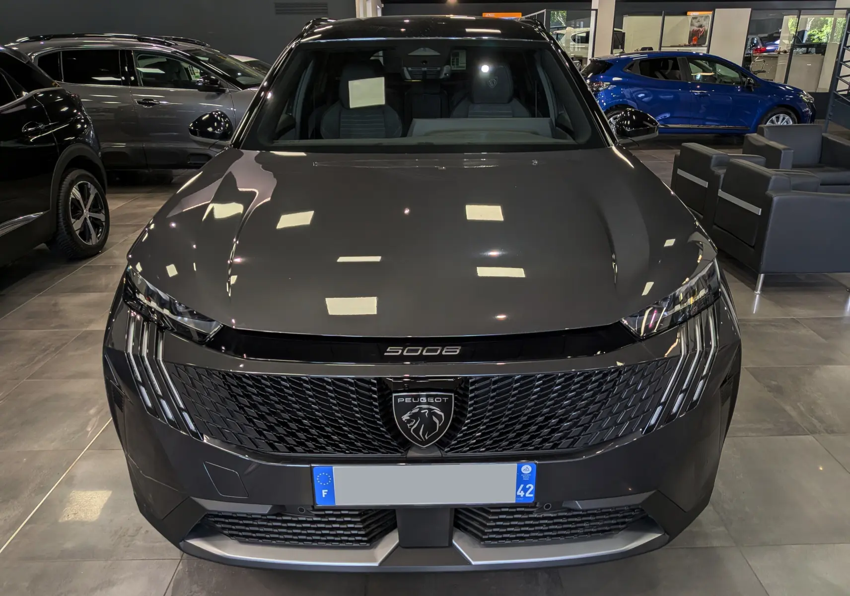 Vue avant d'un Peugeot 5008 Hybrid gris titane avec toit noir, calandre noire et logo lion central visible en showroom.