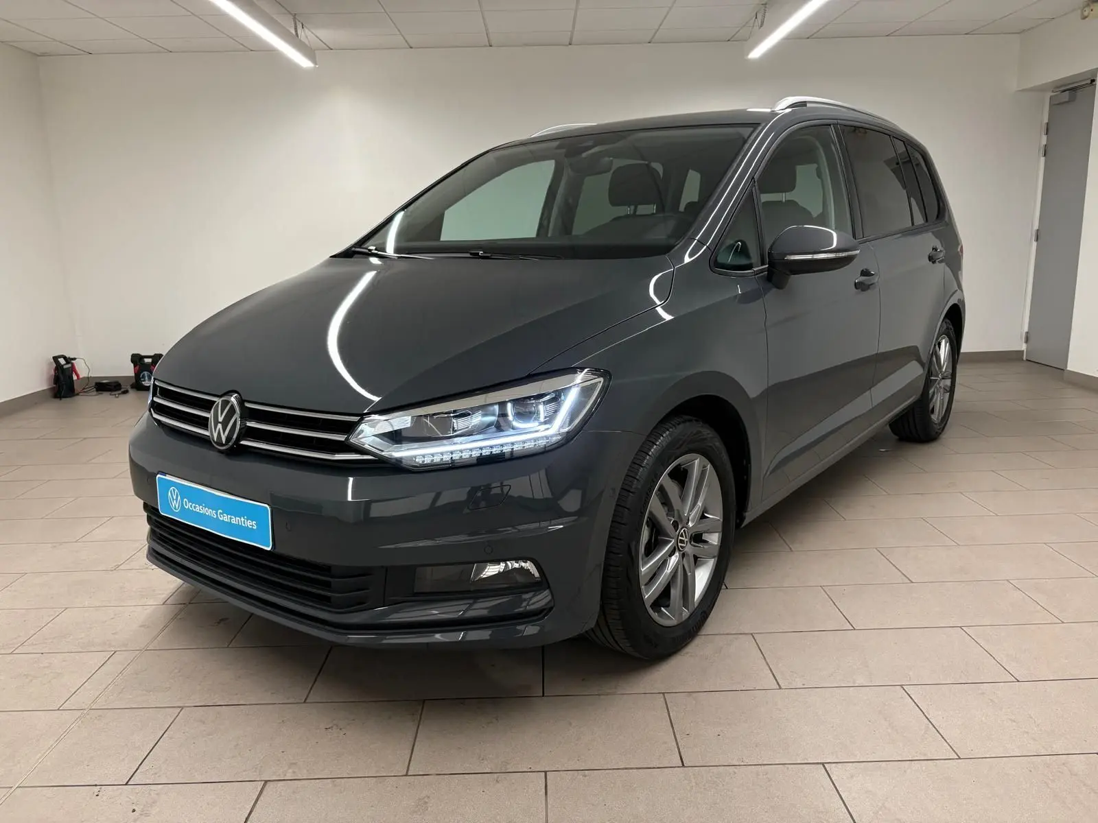 Vue 3/4 avant droit d’un Volkswagen Touran gris dauphin métallisé, avec feux LED allumés en intérieur neutre.
