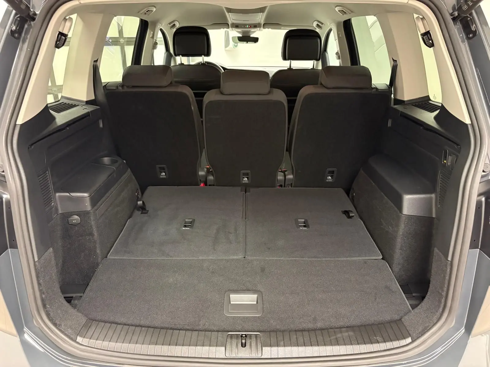 Vue arrière du coffre ouvert du Volkswagen Touran gris dauphin, sièges arrière partiellement rabattus et espace de chargement spacieux.