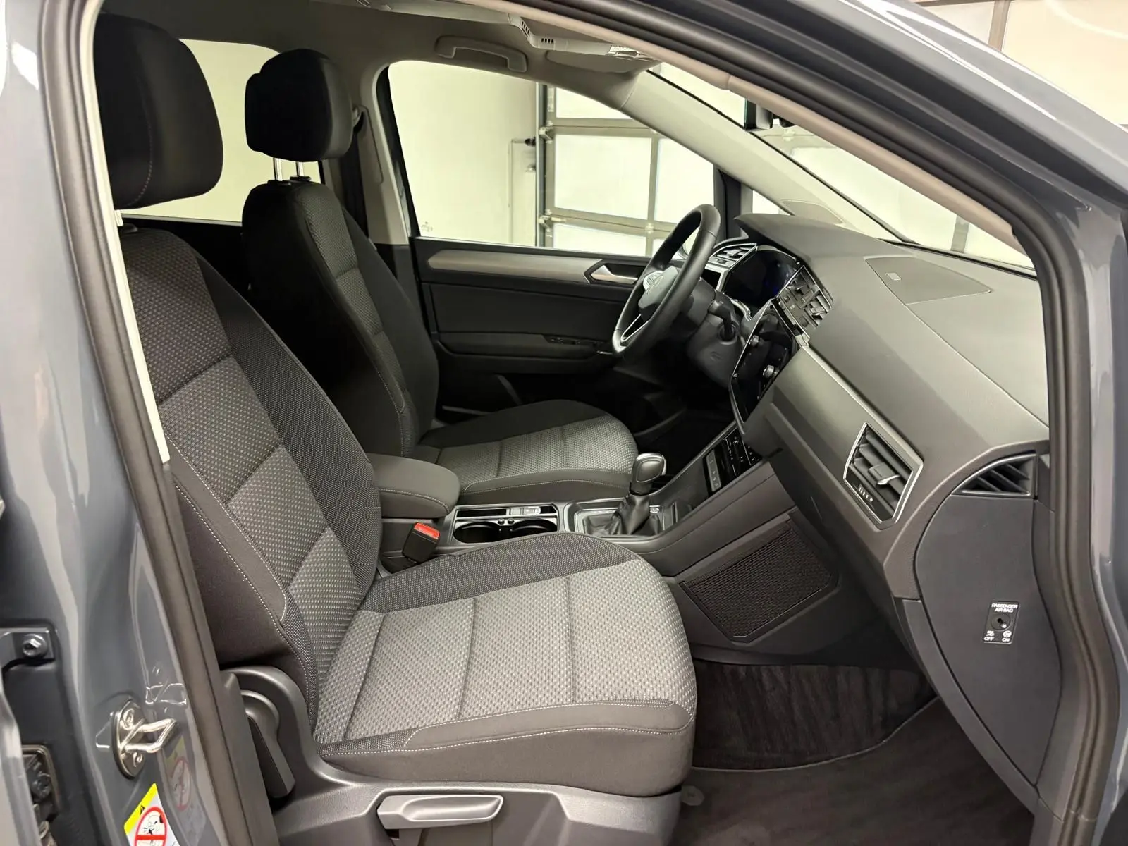 Vue intérieure côté conducteur du Volkswagen Touran gris dauphin, montrant les sièges et le tableau de bord moderne.