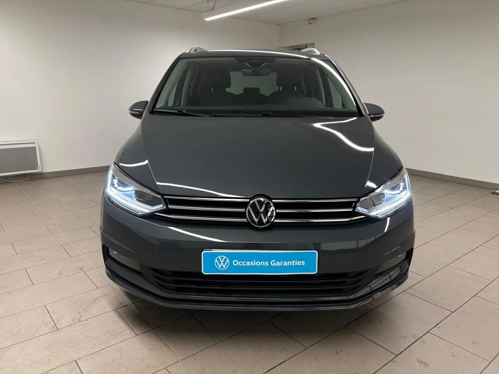 Vue frontale d'un Volkswagen Touran gris dauphin avec phares allumés dans un showroom intérieur.