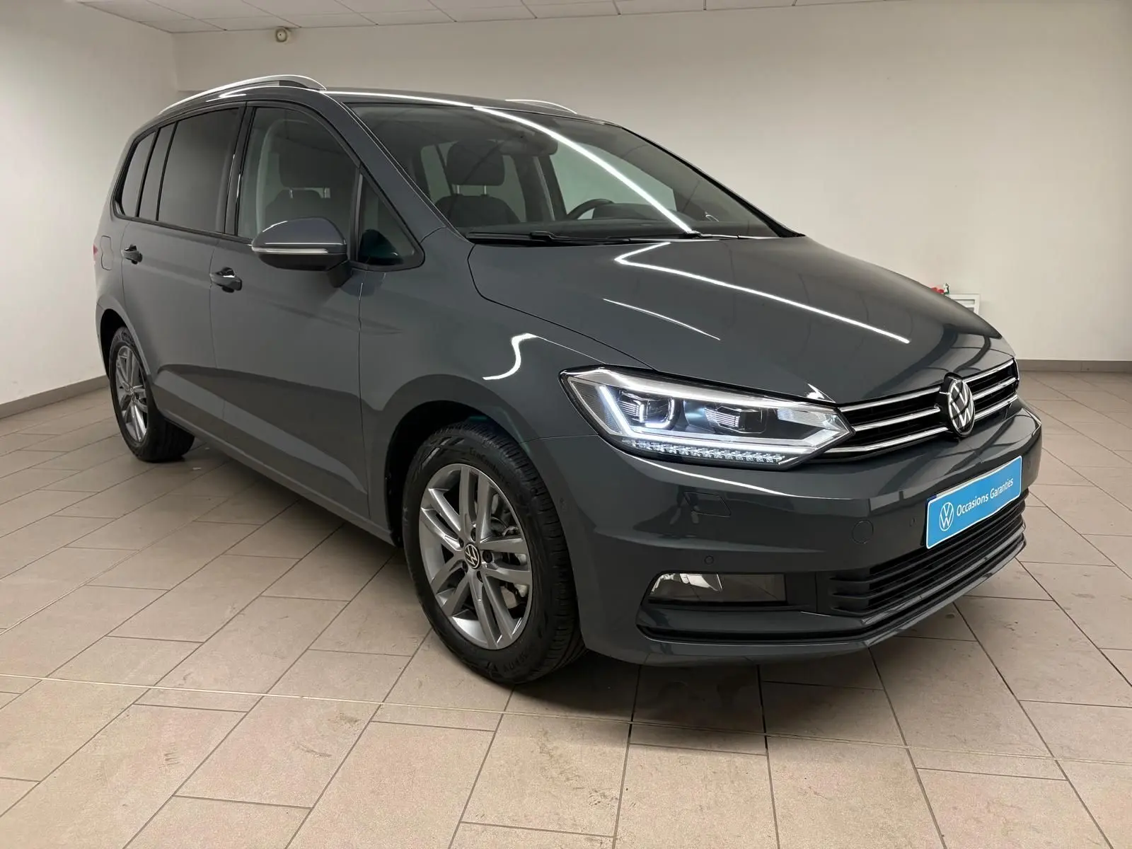 Vue 3/4 avant gauche d’un Volkswagen Touran 2025 gris dauphin avec phares allumés dans un showroom.