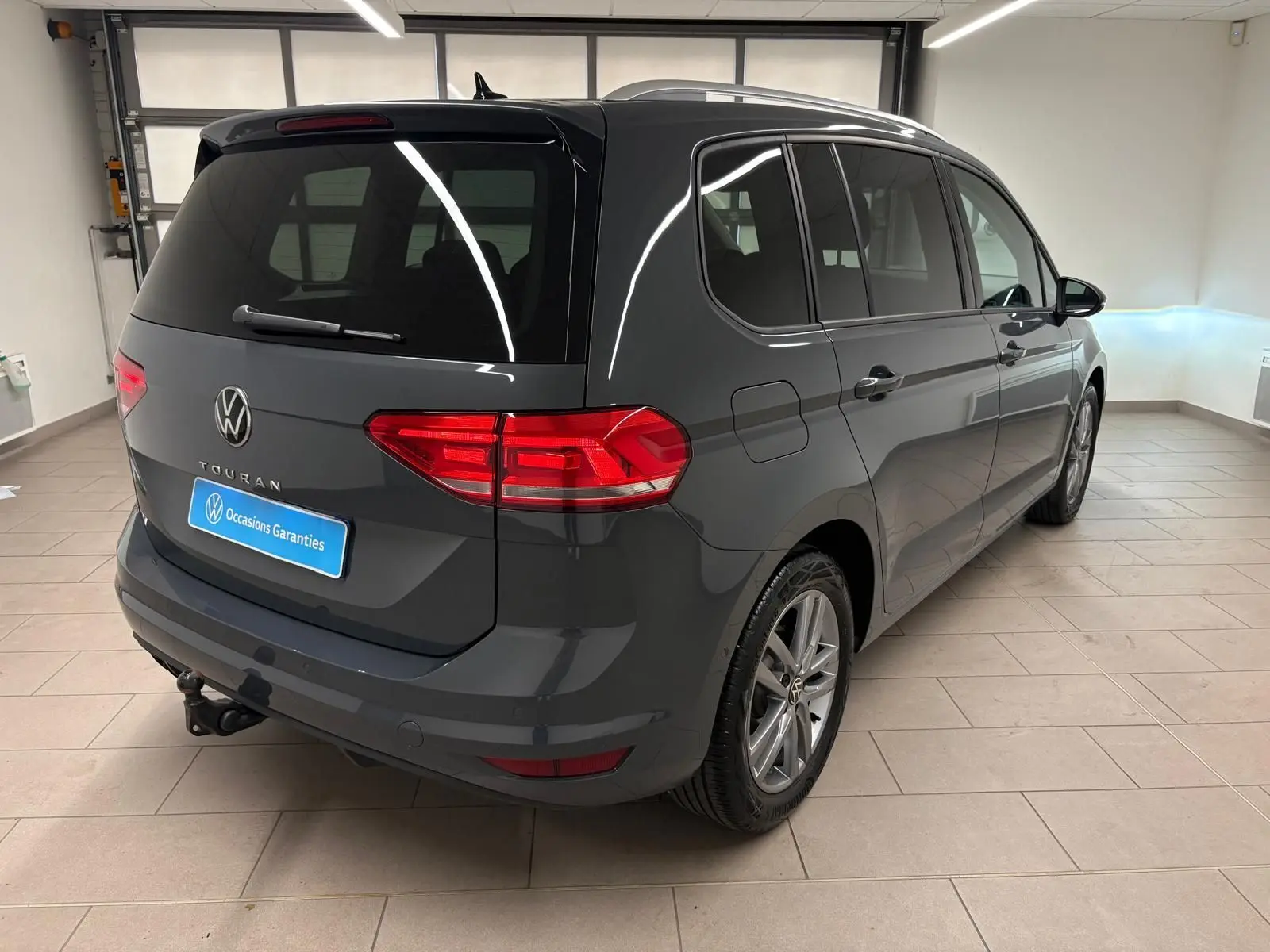 Vue 3/4 arrière droite d’un Volkswagen Touran gris dauphin avec attelage escamotable dans un garage lumineux.